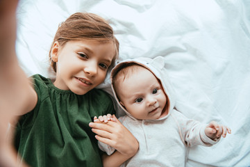 top view of smiling child looking at camera while lying on white bedding near cute baby