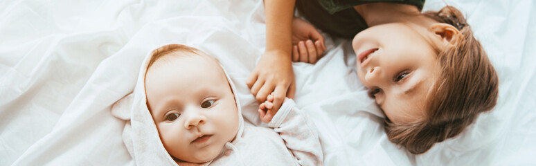 panoramic shot of smiling child holding hand of little sister lying on white bedding