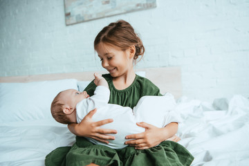 happy child holding cute little sister while sitting on white bedding