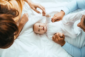 Fototapeten top view of parents touching smiling infant lying on white bedding © LIGHTFIELD STUDIOS