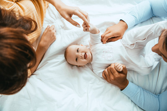 top view of parents touching smiling infant lying on white bedding