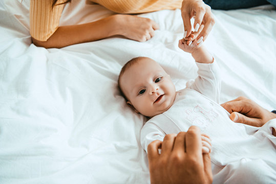 cropped view of mother and dad playing with smiling baby lying on white bedding