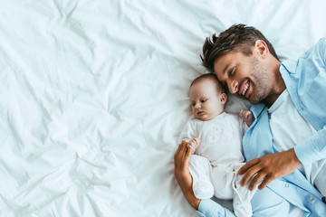 top view of happy father hugging adorable baby while lying on white bedding