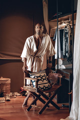 Masculine Arabic businessman in beige silk bathrobe is standing in his walk-in wardrobe, his clothes, shoes and chair around; fashion concept.