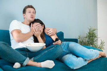 Fototapeta na wymiar young couple beautiful brunette woman and handsome man watching scary movie on the sofa eating pop corn looking frightened he closes her eyes homesters