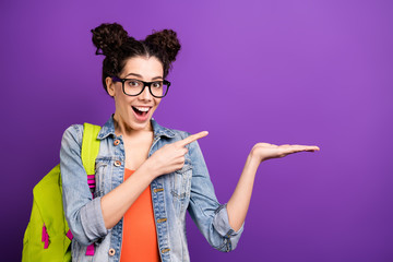 Close up photo of crazy cheerful high school girl hold hand point index finger indicate academic campus option wear denim jeans jacket green rucksack spectacles isolated over purple color background