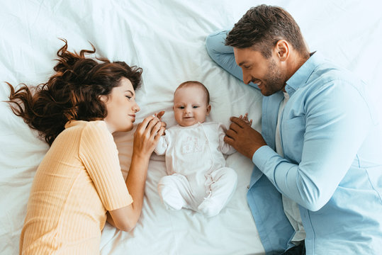 Happy Mom And Dad Gently Touching Adorable Baby While Lying On White Bedding Together