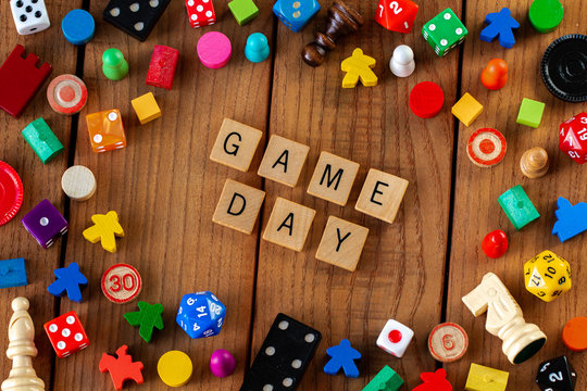 "Game Day" spelled out in wooden letter tiles. Surrounded by dice, cards, and other game pieces on a wooden background