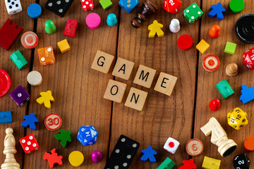 "Game On" spelled out in wooden letter tiles. Surrounded by dice, cards, and other game pieces on a wooden background