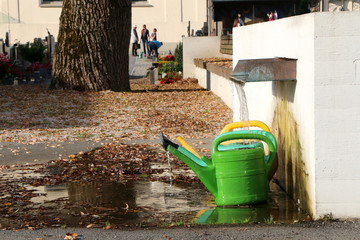 Am Friedhof, Grabpflege, ein Brunnen mit Gießkannen, im unscharfen Hintergrund unkenntliche Personen bei der Grabpflege auf einem Friedhof