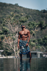 Muscular and handsome shirtless Arabic man in turquoise sarong and sunglasses standing at the...