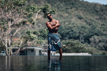 Artistic and handsome shirtless Arabic man in turquoise sarong and sunglasses standing at the poolside in a sunny day, his arms crossed; palms and sky; paradise concept.