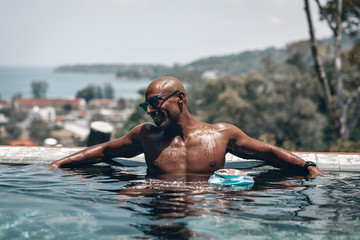 Sensual topless Arabic male in the clear pool in a sunny day, the man is relaxed, he listens to music, floating boombox by his side; the view of the town behind him.