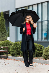 attractive and smiling businesswoman in black coat holding umbrella and paper cup