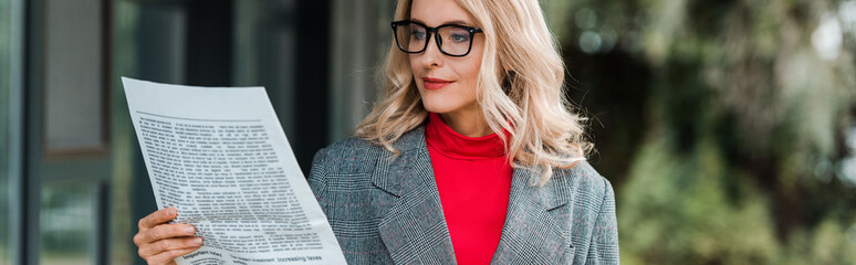 panoramic shot of attractive businesswoman in coat and glasses reading newspaper