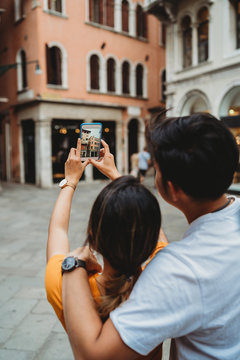 Portrait of young and beautiful Asian couple visit the city of Venice, Italy - Millennials on their honeymoon take a picture with a smart phone at buildings town square - The man embraces woman