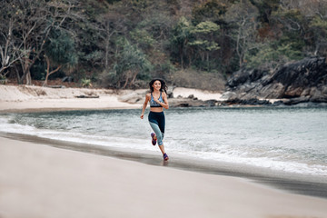 A cute fit woman jogs  across the sandy beach by the ocean in Turkey. Rocks and trees behind.