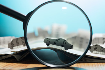 Magnifying glass in front of an open newspaper with paper cars. Concept of car rent, search, purchase and cargo transportation.