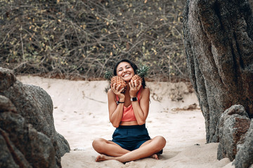 Hilarious young girl in lotus pose at the beach holds two pineapples by her chin.