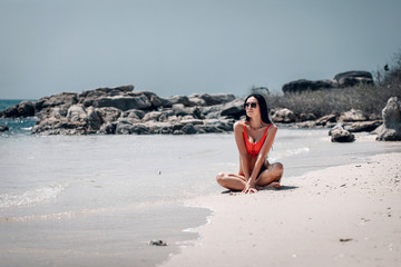 Beautiful tanned lady in red bikini and sunglasses sits on the beach on the seaside, relaxation concept.