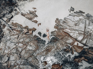 Top view of the fit woman lying on coastal rocks by turquoise ocean waters; drone shot.