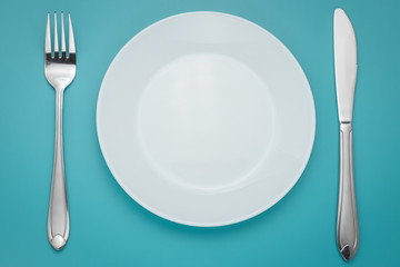 White empty plate with fork and knife on blue background. Turquoise backdrop. Laying, table appointments.
