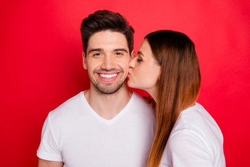 Photo of redhair positive cute nice pretty couple of married people with wife in white t-shirt kissing her husband cheek smiling toothily showing their tender love isolated bright red color background - obrazy, fototapety, plakaty