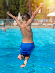 Cute active European boy in blue swimming shorts having fun in hotel’s swimming pool, he is making fantastic jump, his hands open wide.