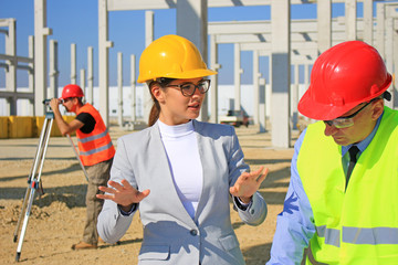 Female architect and construction engineer in hardhats talking about the project on construction site, behind them construction worker with measuring device, teamwork