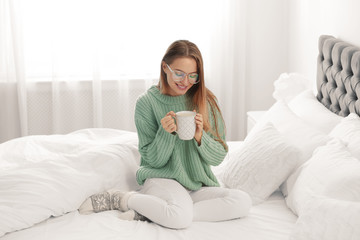 Young woman in warm sweater with hot drink on bed at home