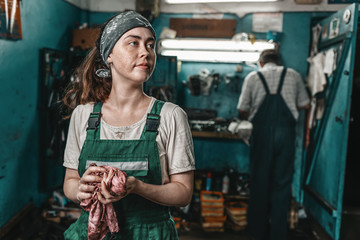 The concept of women's equality and feminism. A woman in a green work uniform wipes her hands with a rag. In the background, a man is working at a machine. Factory and workshops