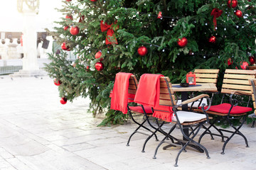 Street cafe under Christmas tree with red ornaments outdoor. Wooden tables and chairs with warm plaid at winter market in old italian town. Cozy festive and New Year atmosphere in Europe