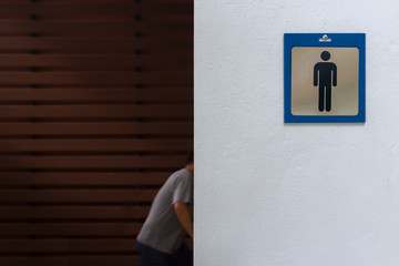Blue frame of male symbol stick by screw nail head on rough white cement wall. Man customer ware gray t-shirt walking in men toilet with blurred dark brown plank wood s and sun lighting background.