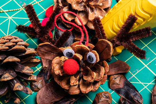 A Macro image of various pine cone reindeer being created.  The pine cones are on a green cutting mat and have bright red pom-pom noses and googly eyes.