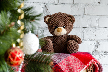charming brown Teddy bear sitting on a red blanket near the Christmas tree decorated with toys and balls and waiting for his mistress, a gift for the child. On a light background of white brick wall