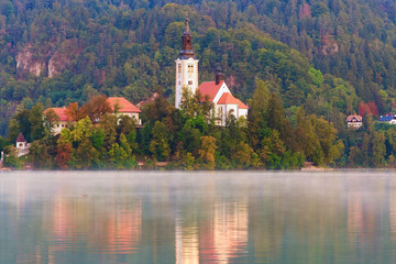 Fototapeta na wymiar Picturesque morning view of Bled Lake and Pilgrimage Church of the Assumption of Maria on small island against autumn forest. Morning fog on the water surface. Travel and tourism concept. Slovenia