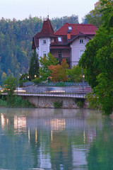 Naklejka na ściany i meble Scenic morning view of ancient traditional house near Bled lake. Morning fog on the water surface and fall colorful trees background. Bled Lake, the Upper Carniolan region of northwestern Slovenia
