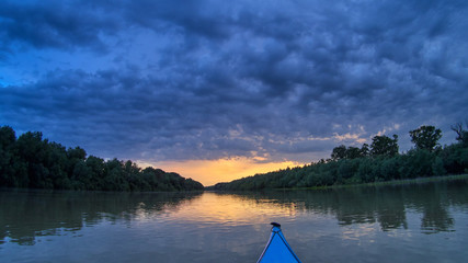 Bow of blue kayak. Dramatic sunset over Danube river with forest along riverside