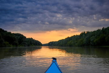 Bow of blue kayak. Dramatic sunset over Danube river with forest along riverside