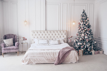 Decorated Christmas tree in white classic bedroom interior with New Year's holiday bouquet in a vase, a gently pink gift present box on the bedside glass table and classic armchair in lavender colors.