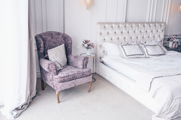 White classic bedroom interior with New Year's holiday bouquet in a vase, a gently pink gift present box on the bedside glass table and classic armchair in lavender colors
