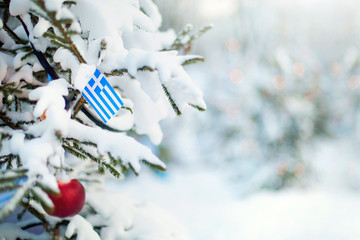 Christmas Greece. Xmas tree covered with snow, decorations and Greek flag. Snowy forest background in winter. Christmas greeting card. - 299390058