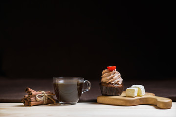 Transparent cup with coffee, cinnamon and cupcake on a wooden cutting board. Dark brown background, light table