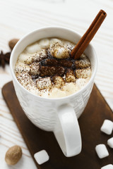 White mug with hot chocolate and marshmallows with cinnamon stick on a wooden table. Hot winter drink, top view.