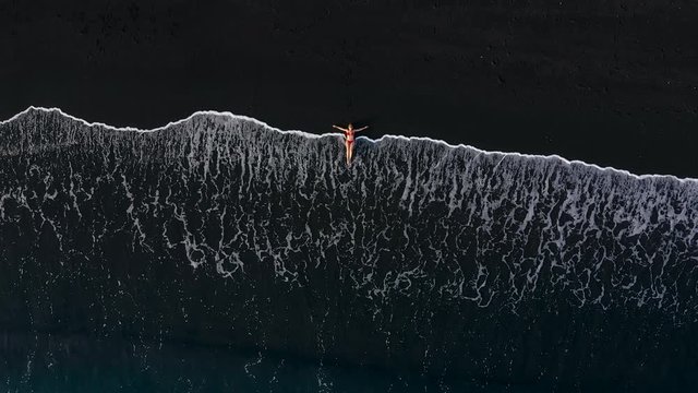 Top view of a woman in a red swimsuit lying on a black beach on the surf line. Coast of the island of Tenerife, Canary Islands, Spain.
