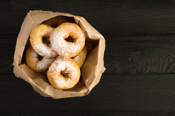 Homemade donuts with powdered sugar in the paper package  on the black wooden background. Top view....