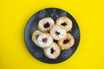 Homemade donuts with powdered sugar in the black plate in the center of the yellow background. Top view. Copy space.