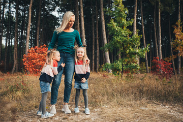 Naklejka na ściany i meble Blonde mother and her twin daughters are walking in the autumn forest among red trees.