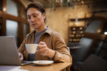 Mature woman drinking coffee and working online on laptop computer at the table in cafe