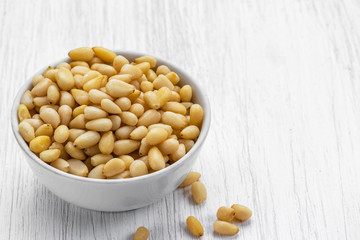 Pine nuts lie in a white bowl on a white wooden surface, side view from above